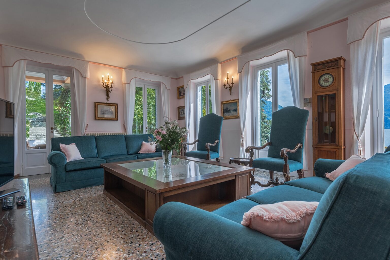 Cozy living room featuring teal sofas, a wooden coffee table, and large windows allowing natural light. Decorated with plants and artwork, the space combines comfort and elegance. | Only on SopranoVillas