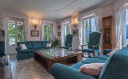 Cozy living room featuring teal sofas, a wooden coffee table, and large windows allowing natural light. Decorated with plants and artwork, the space combines comfort and elegance.