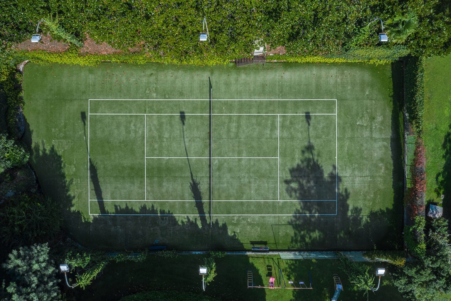 Aerial view of a well-maintained tennis court surrounded by lush greenery, showcasing clear lines and shadows from surrounding trees. | Only on SopranoVillas