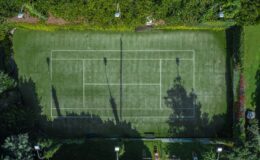 Aerial view of a well-maintained tennis court surrounded by lush greenery, showcasing clear lines and shadows from surrounding trees.