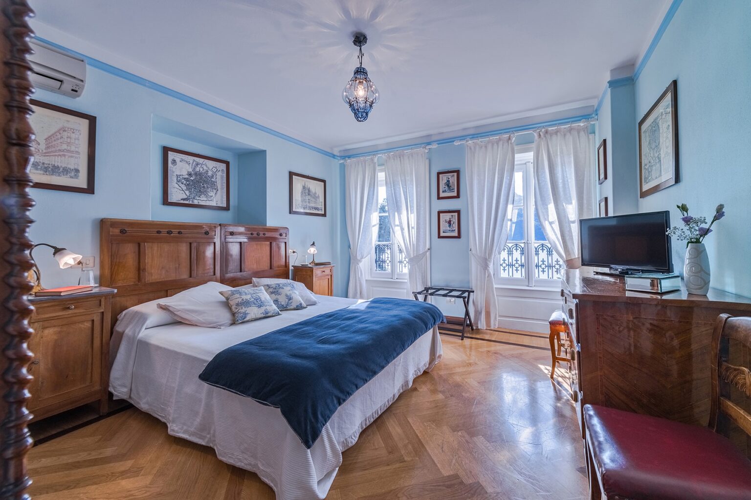 Cozy bedroom with blue walls, hardwood flooring, and a large window letting in natural light. Features a double bed with white linens and a navy throw, antique wooden furniture, decorative artwork, and a television. Ideal for a relaxing stay. | Only on SopranoVillas