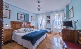 Cozy bedroom with blue walls, hardwood flooring, and a large window letting in natural light. Features a double bed with white linens and a navy throw, antique wooden furniture, decorative artwork, and a television. Ideal for a relaxing stay.