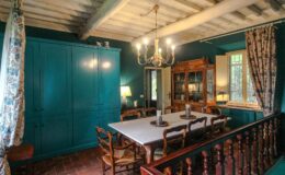 Charming dining room featuring a large wooden table surrounded by rustic chairs, teal walls, and elegant chandeliers, highlighting a cozy and inviting atmosphere.