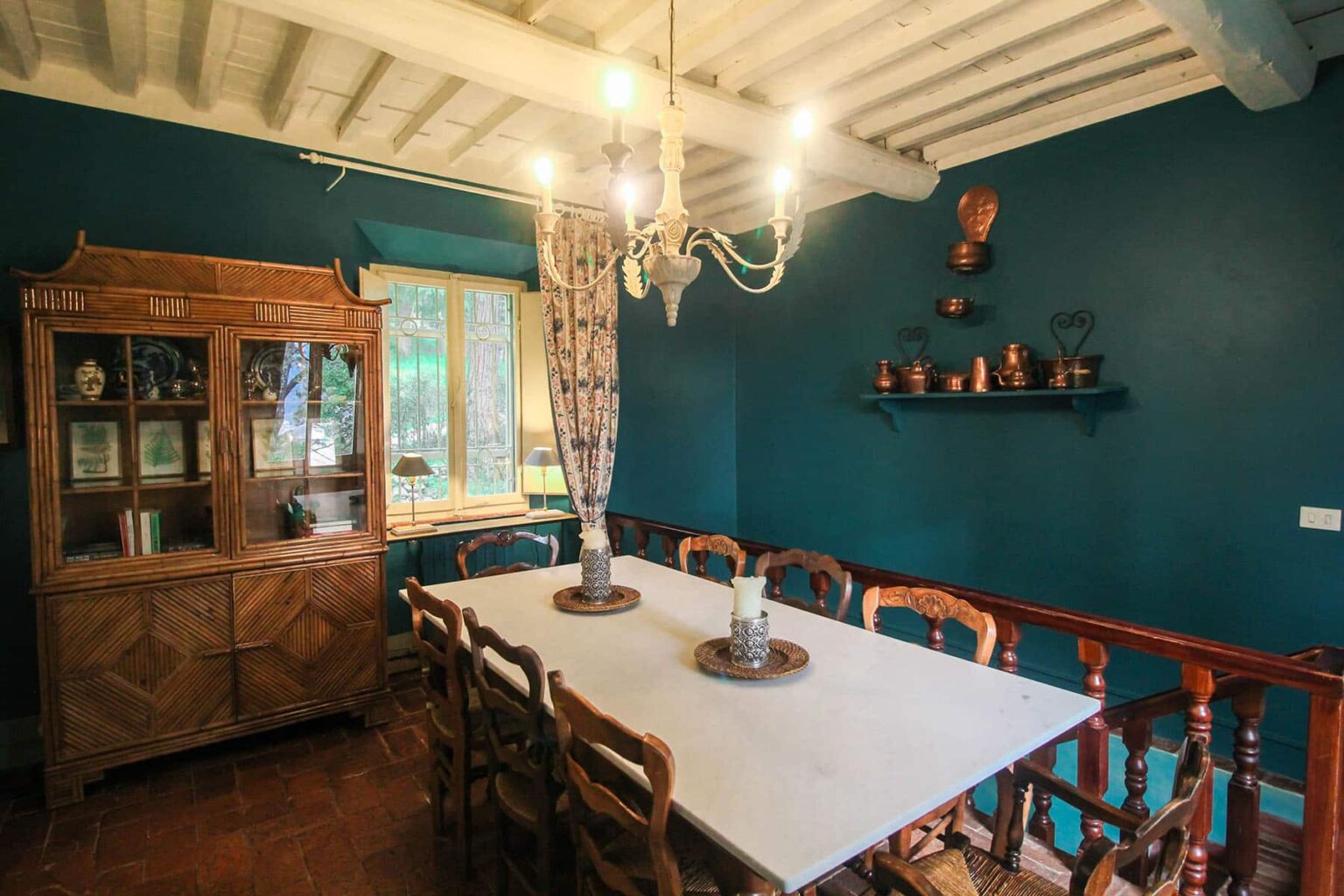 Cozy dining room featuring a large white table surrounded by wooden chairs, a vintage wooden cabinet, and a chandelier, all set against a rich teal wall with decorative shelves. | Only on SopranoVillas