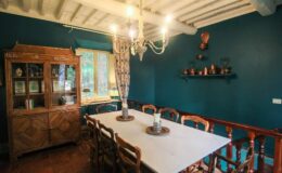 Cozy dining room featuring a large white table surrounded by wooden chairs, a vintage wooden cabinet, and a chandelier, all set against a rich teal wall with decorative shelves.