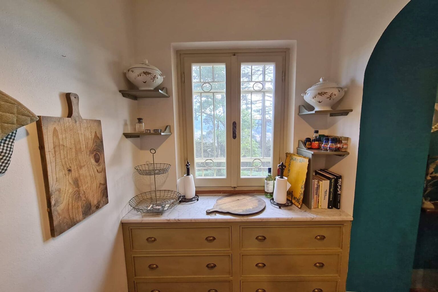 Cozy kitchen corner featuring a wooden dresser with a marble top, decorative shelving, a window with a view, and various kitchen items including books, dishes, and utensils. | Only on SopranoVillas