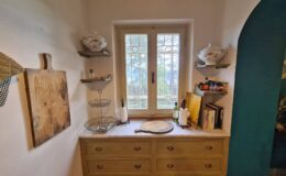 Cozy kitchen corner featuring a wooden dresser with a marble top, decorative shelving, a window with a view, and various kitchen items including books, dishes, and utensils.