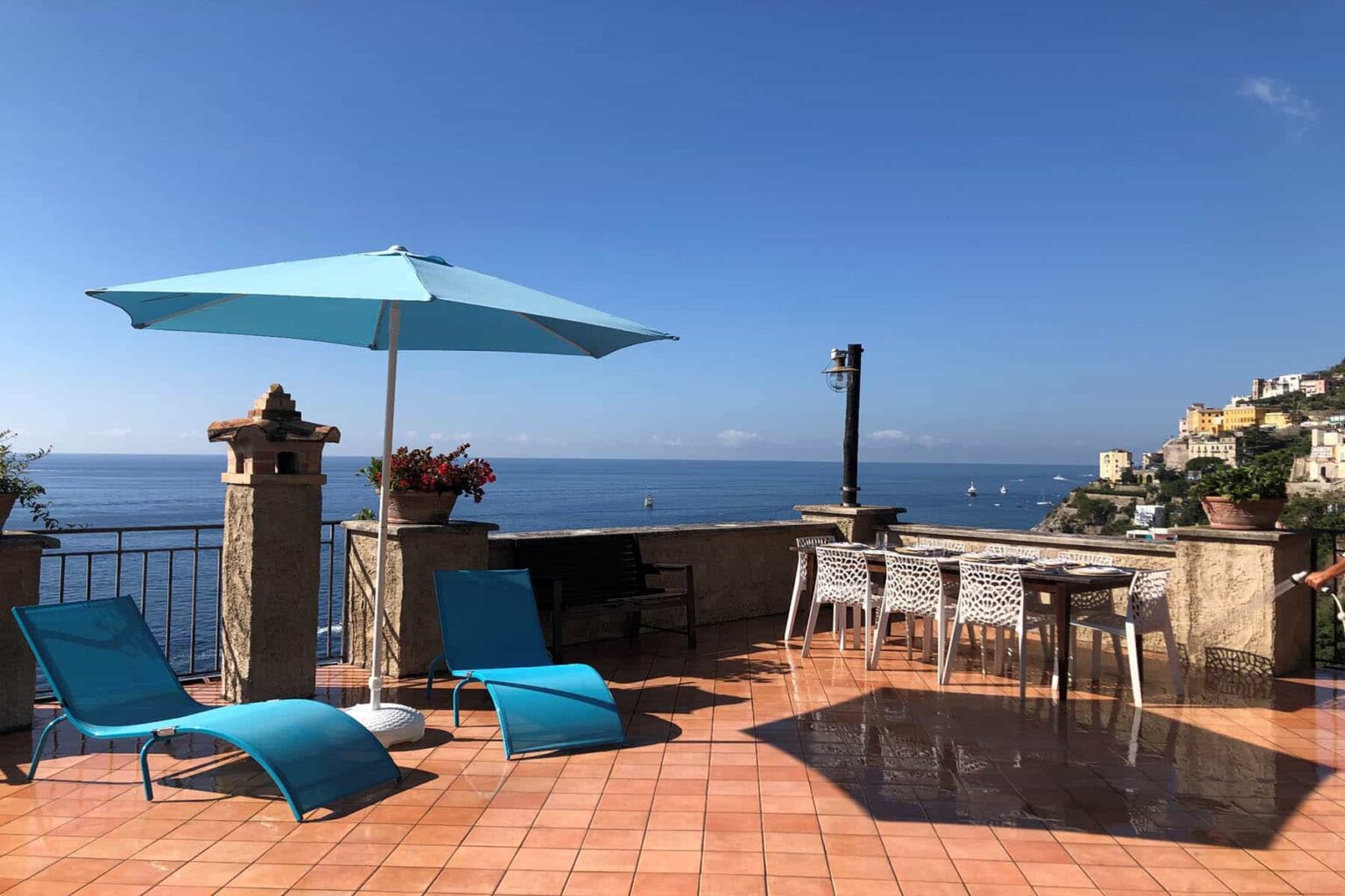 Outdoor terrace overlooking the sea, featuring blue lounge chairs, a large turquoise umbrella, and a dining table with chairs, set against a clear blue sky and coastal scenery. Perfect for relaxation and enjoying stunning views. | Only on SopranoVillas