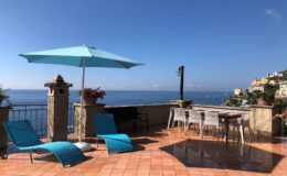 Outdoor terrace overlooking the sea, featuring blue lounge chairs, a large turquoise umbrella, and a dining table with chairs, set against a clear blue sky and coastal scenery. Perfect for relaxation and enjoying stunning views.