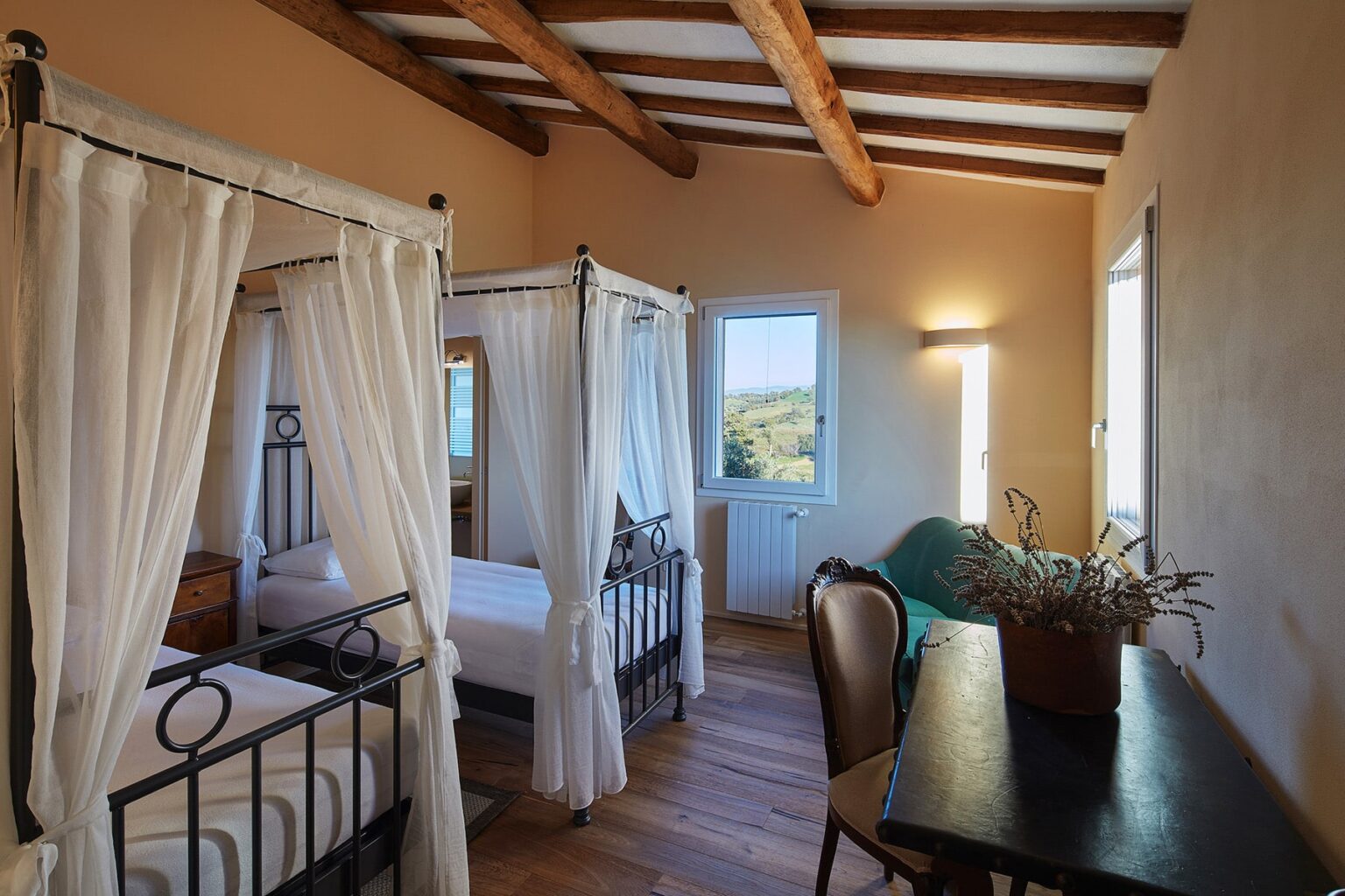 Cozy bedroom featuring two canopy beds with white drapes, wooden ceiling beams, and a desk with a potted plant, offering a serene view through large windows. | Only on SopranoVillas