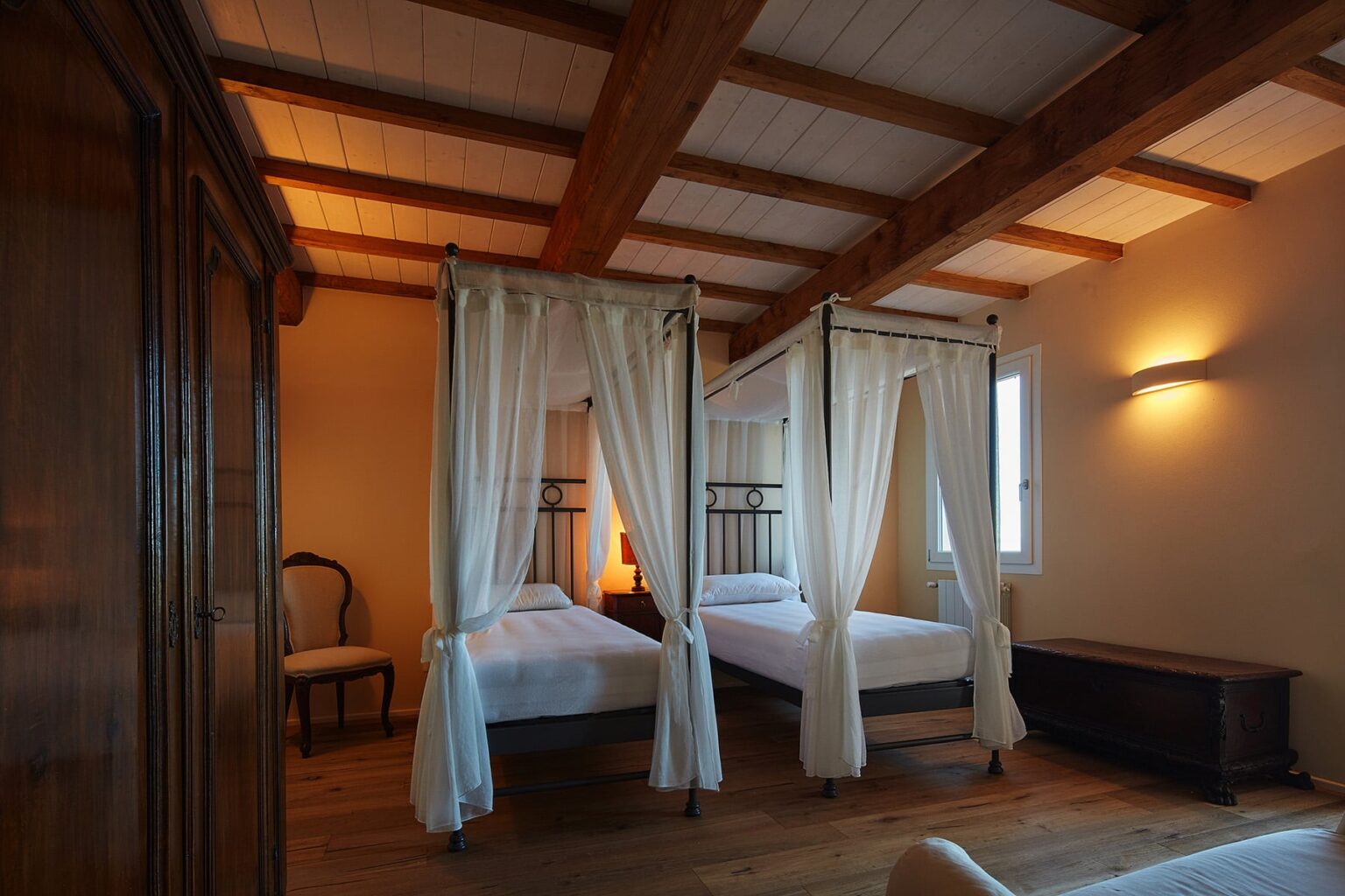 Cozy bedroom featuring two four-poster beds with white drapes, wooden beams on the ceiling, and warm lighting, creating a welcoming and elegant atmosphere. | Only on SopranoVillas