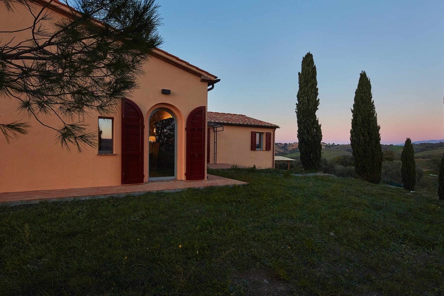 Charming Tuscan villa at dusk, featuring a warm exterior with red shutters, surrounded by lush green grass and tall cypress trees, set against a serene landscape. | Only on SopranoVillas