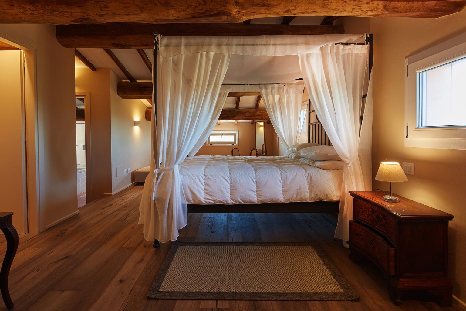 Cozy bedroom featuring a four-poster bed with sheer white curtains, wooden beams, and warm lighting, complemented by a bedside table and a soft area rug. Ideal for a relaxing retreat. | Only on SopranoVillas