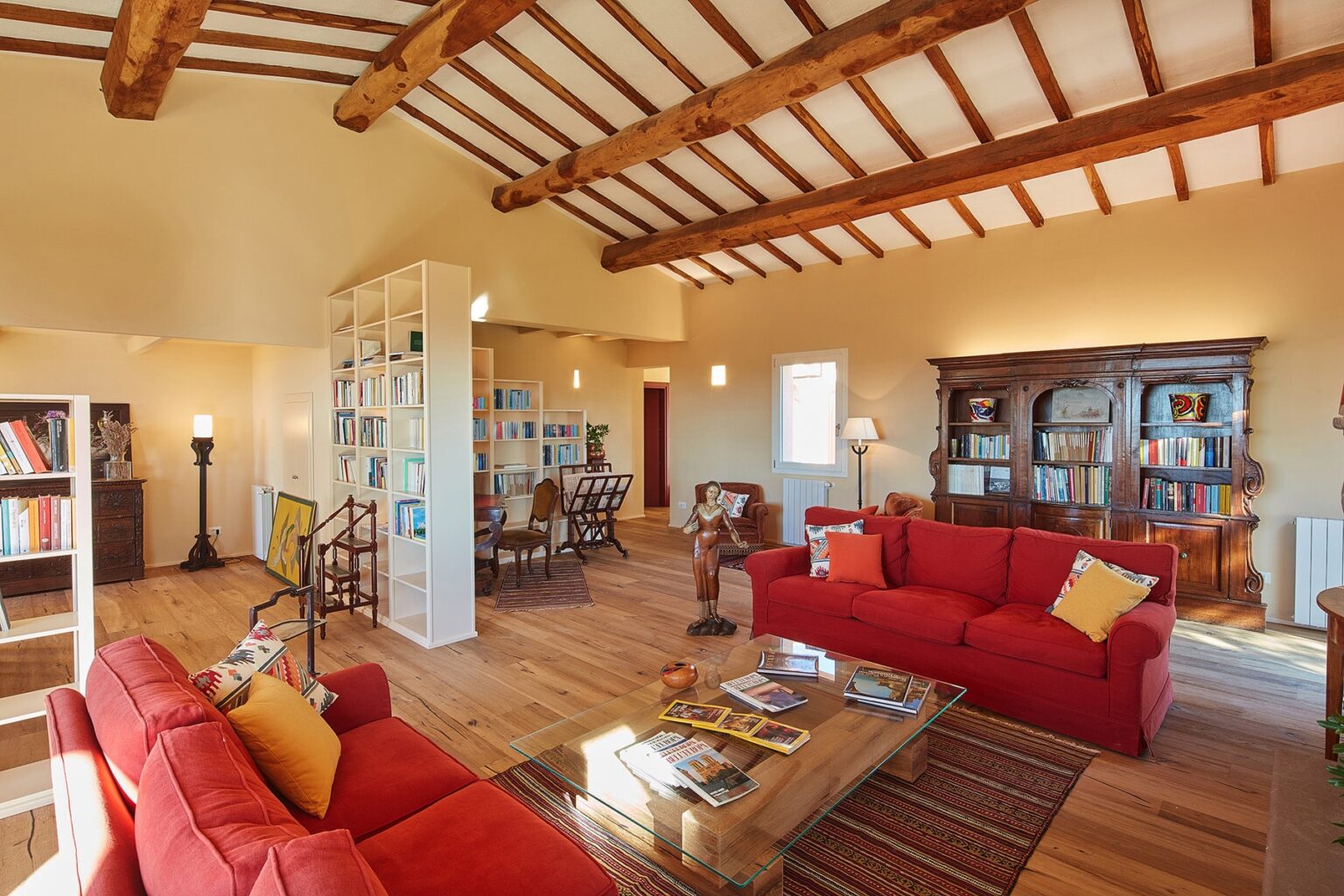 Spacious living room with high wooden beams, featuring red sofas, a wooden coffee table, and a bookshelf. Natural light illuminates the warm, inviting space, which includes a dining area in the background. | Only on SopranoVillas