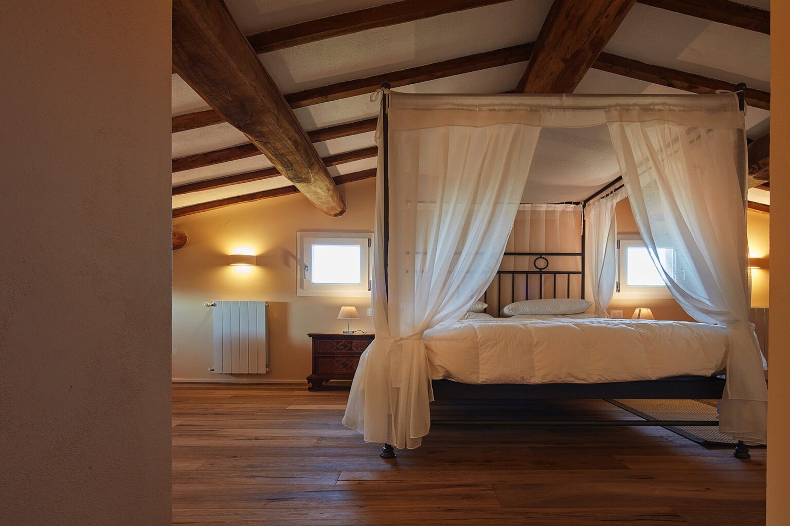 Cozy bedroom featuring a four-poster bed with sheer drapes, wooden beams on the ceiling, soft lighting, and warm wooden flooring, creating a tranquil and inviting atmosphere. | Only on SopranoVillas