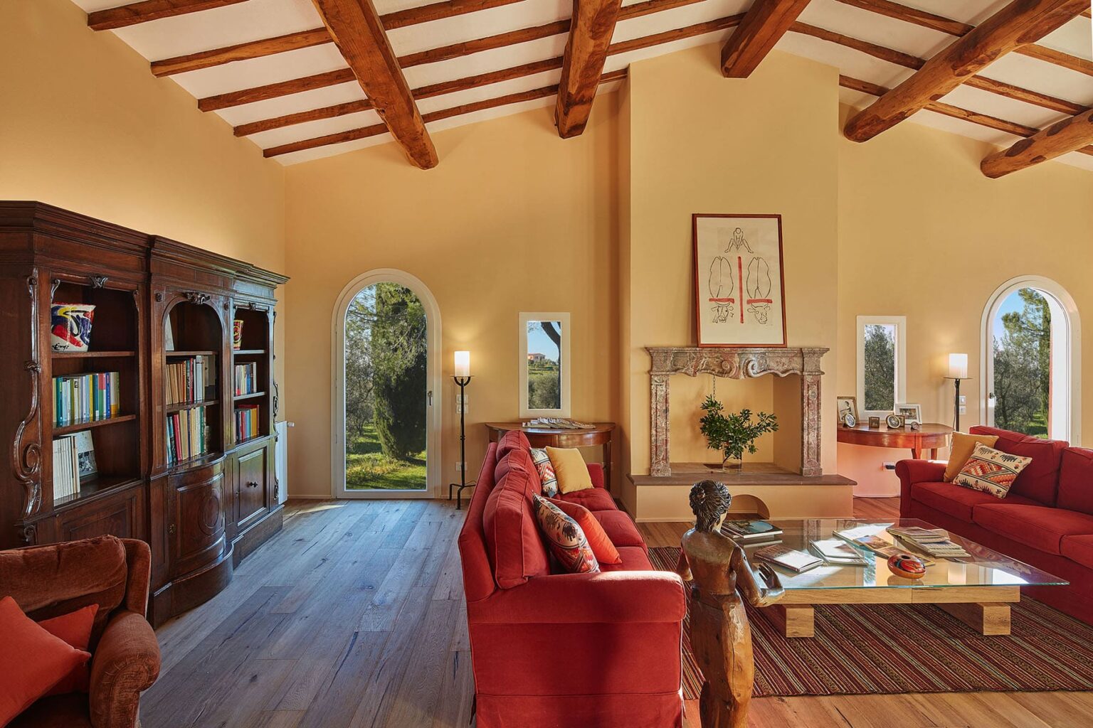Cozy living room featuring red sofas, a wooden coffee table, and a stone fireplace, with large windows allowing natural light and views of the outdoors. The room has exposed wooden beams and a bookshelf filled with books, creating a warm and inviting atmosphere. | Only on SopranoVillas