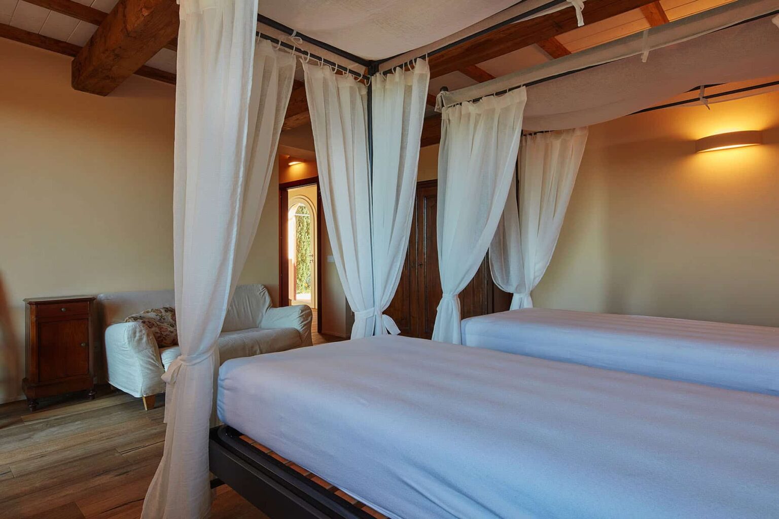 Cozy bedroom featuring two twin beds with white bedding, draped with sheer curtains, wooden beams on the ceiling, and a stylish armchair in the background, offering a serene and inviting atmosphere. | Only on SopranoVillas