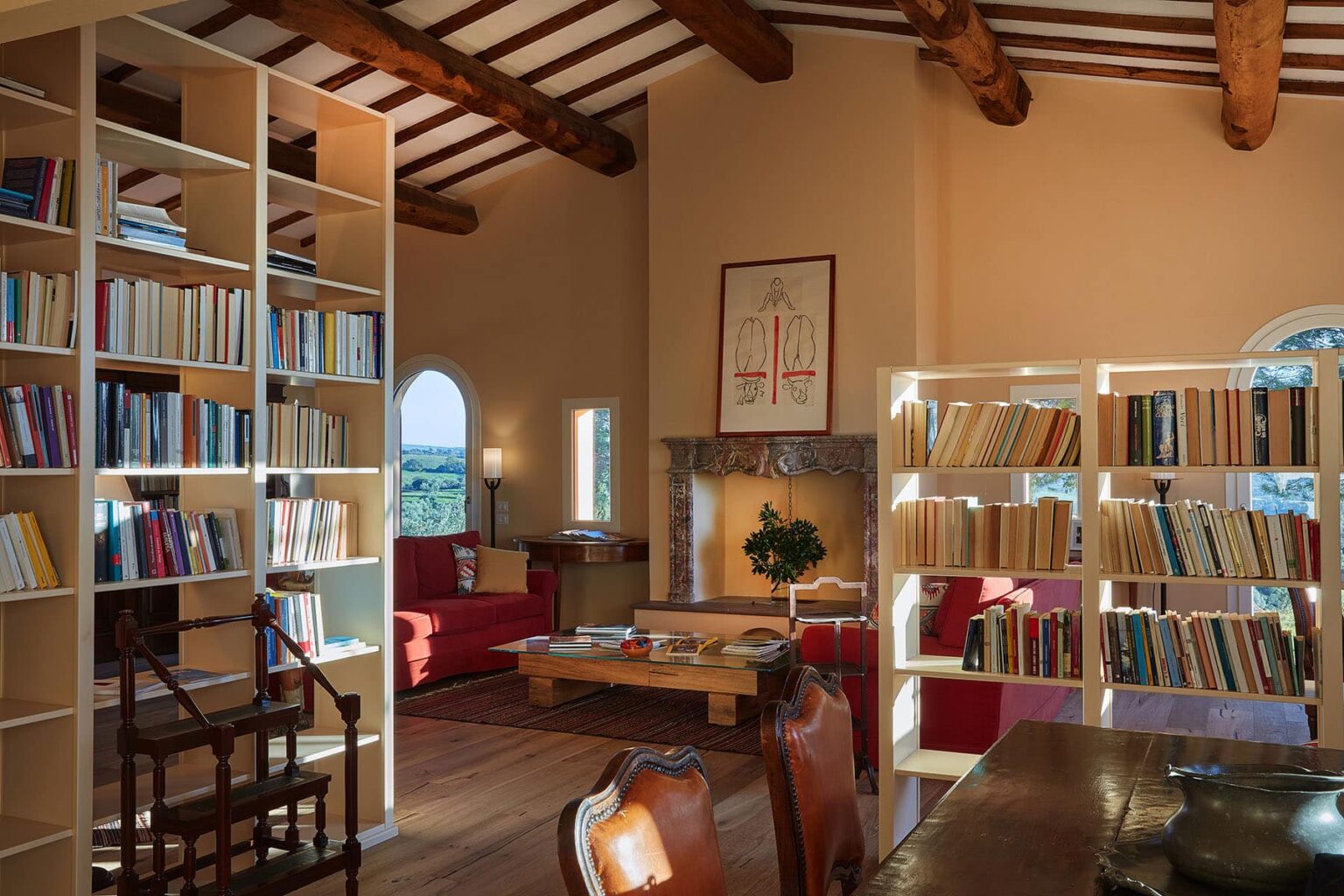 Cozy reading room featuring tall bookshelves filled with books, a wooden table, and comfortable seating. The space is illuminated by natural light from arched windows, highlighting the rustic wooden beams and warm color palette. Ideal for relaxation and study. | Only on SopranoVillas