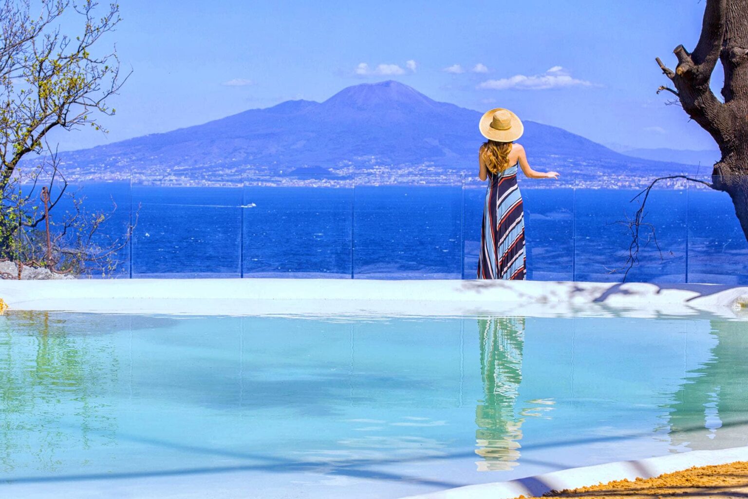 Woman in a striped dress and sun hat stands by a serene pool overlooking the blue waters and Mount Vesuvius in the background, creating a picturesque vacation scene. | Only on SopranoVillas