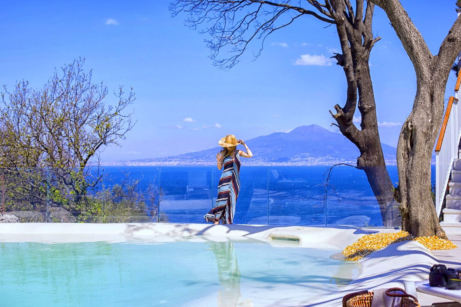 Woman in a stylish dress and sunhat standing by a pool with a stunning ocean view, featuring a clear blue sky and distant mountains. Ideal for travel and leisure content. | Only on SopranoVillas