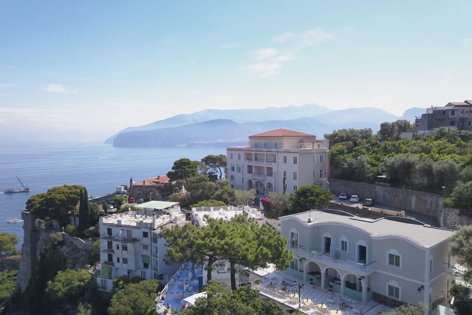Aerial view of coastal villas and lush greenery overlooking the sea, with mountains in the background, showcasing the scenic beauty of a Mediterranean destination. | Only on SopranoVillas