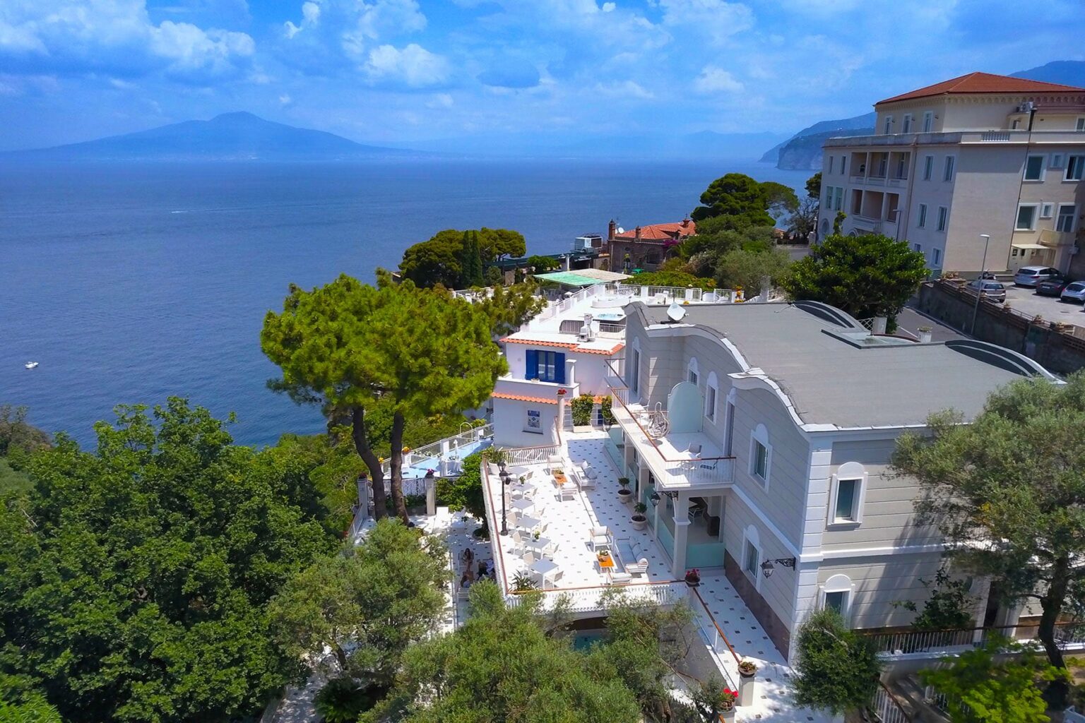 Aerial view of a luxurious coastal villa overlooking the ocean, surrounded by greenery and with distant mountains in the background. The property features a spacious terrace and modern architecture, ideal for vacation rentals or luxury getaways. | Only on SopranoVillas
