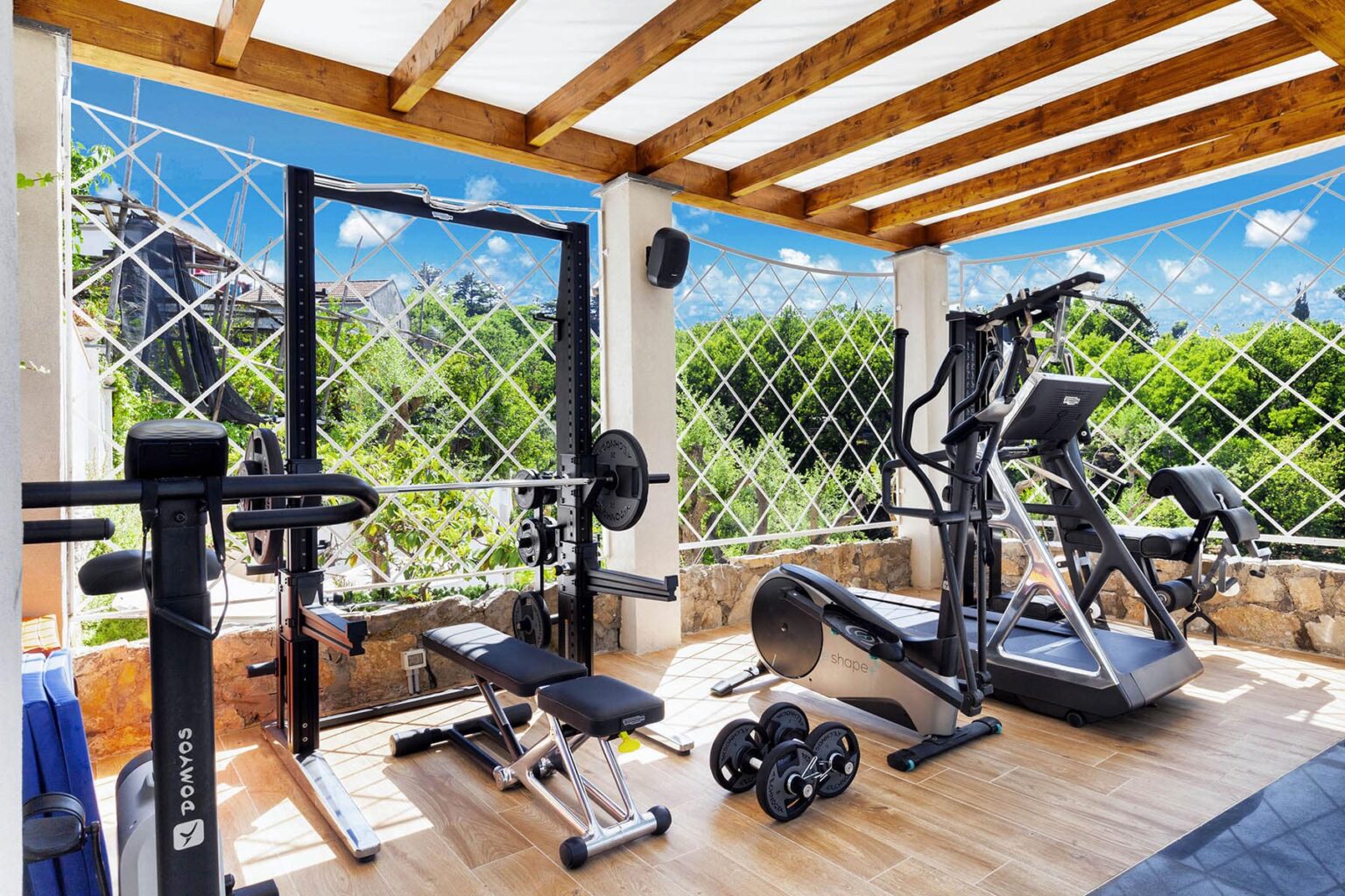 Home gym with various fitness equipment, including a weight bench, barbell, and elliptical machine, set in a bright, airy space surrounded by greenery and natural light. | Only on SopranoVillas