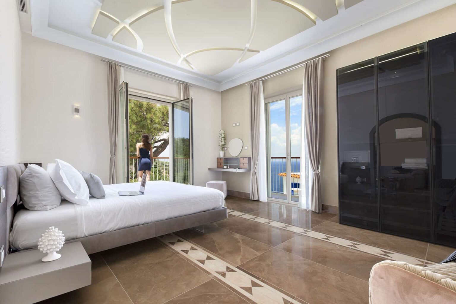 Modern bedroom with large windows and a scenic view, featuring a stylish bed, elegant decor, and a person standing by the door. The room has marble flooring and contemporary furnishings, creating a luxurious ambiance. | Only on SopranoVillas