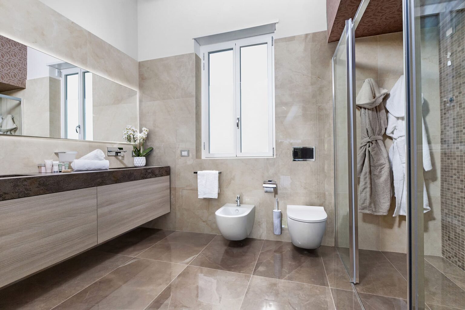 Modern bathroom featuring elegant beige tiles, dual washbasins, a bidet, and a glass shower enclosure, with natural light streaming through a window. | Only on SopranoVillas