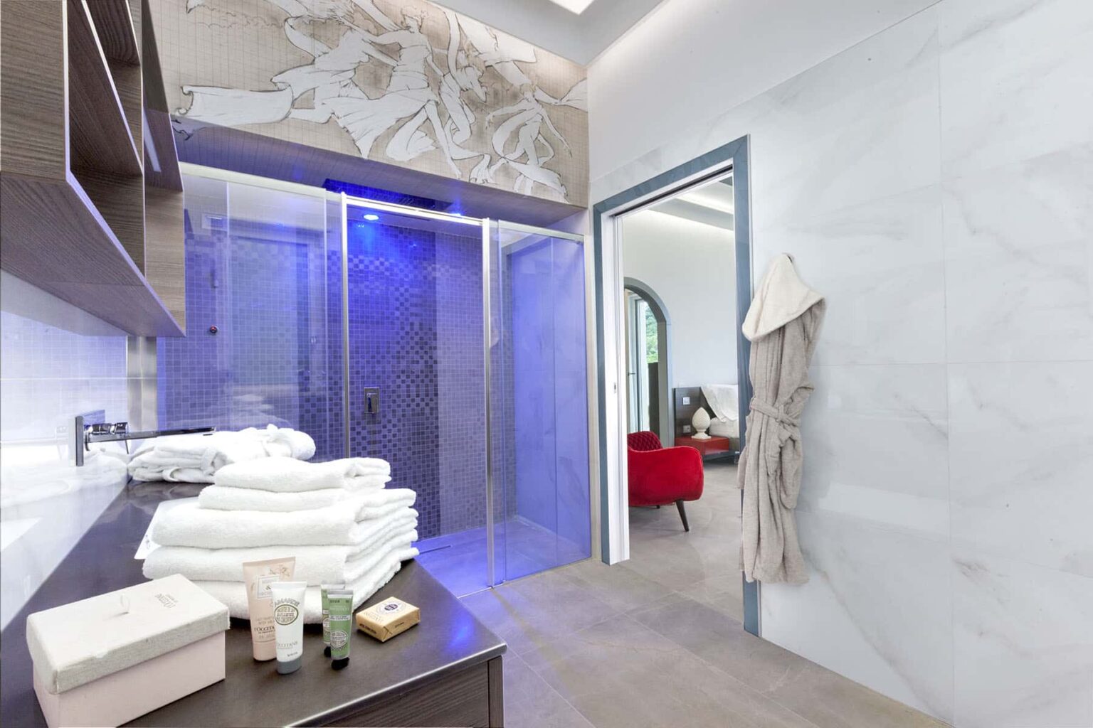 Modern bathroom interior featuring a glass shower, neatly stacked white towels, and elegant decor. The space includes a stylish robe hanging on the wall and a glimpse of a cozy living area in the background. | Only on SopranoVillas