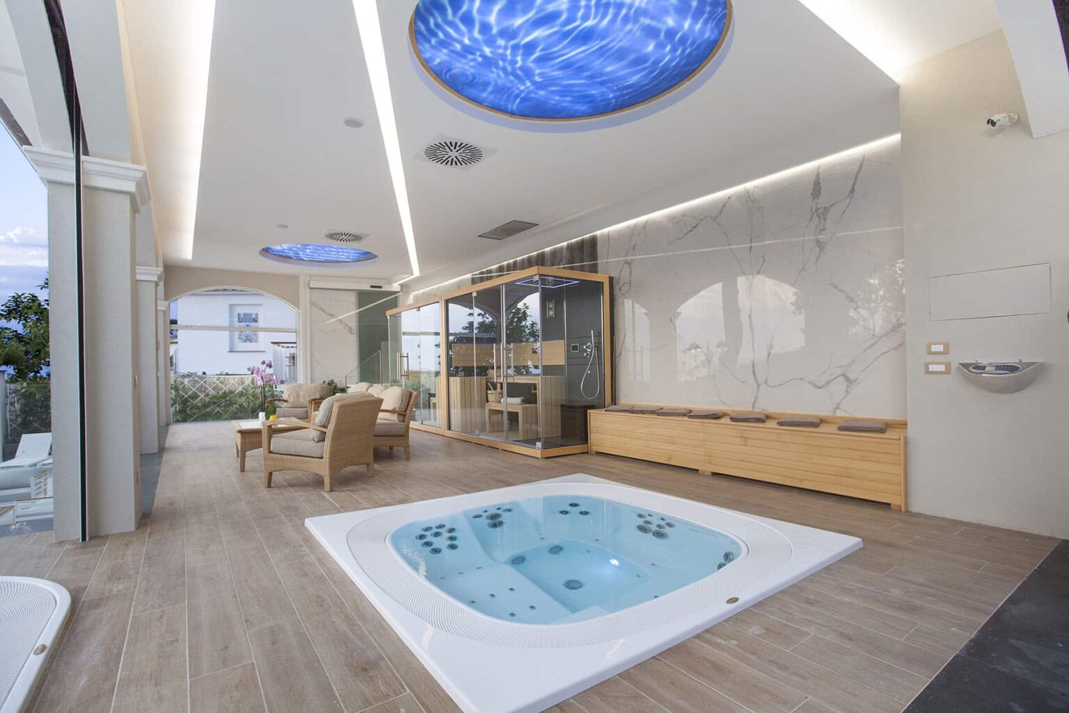 Modern indoor spa area featuring a luxurious hot tub, wooden accents, and elegant ceiling designs with blue lighting. The space is designed for relaxation and wellness, showcasing a serene atmosphere with natural light and stylish furnishings. | Only on SopranoVillas