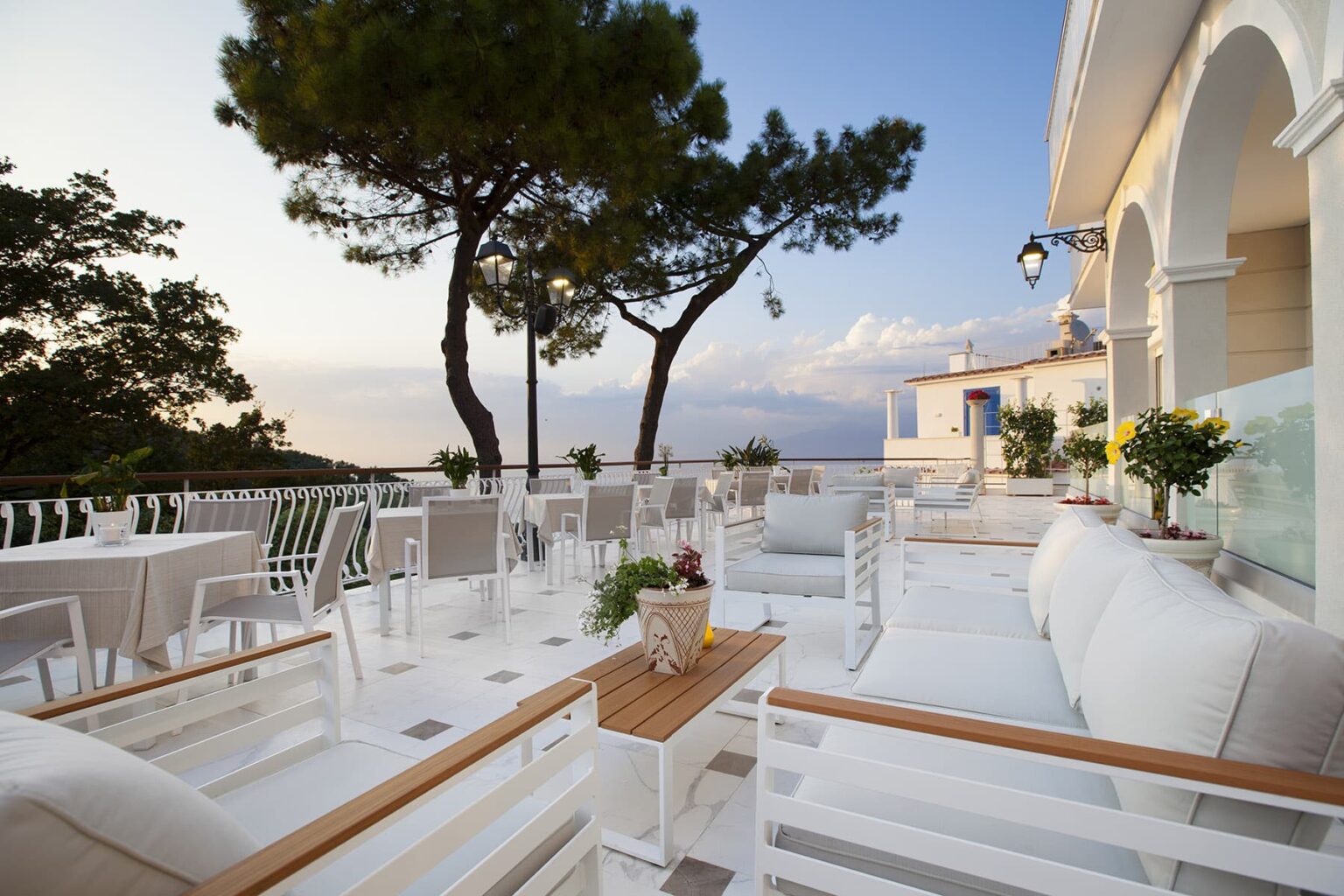 Outdoor terrace of a stylish restaurant featuring white furniture, wooden accents, and lush greenery, with a scenic view of the sky and distant mountains. Ideal for dining and relaxation. | Only on SopranoVillas