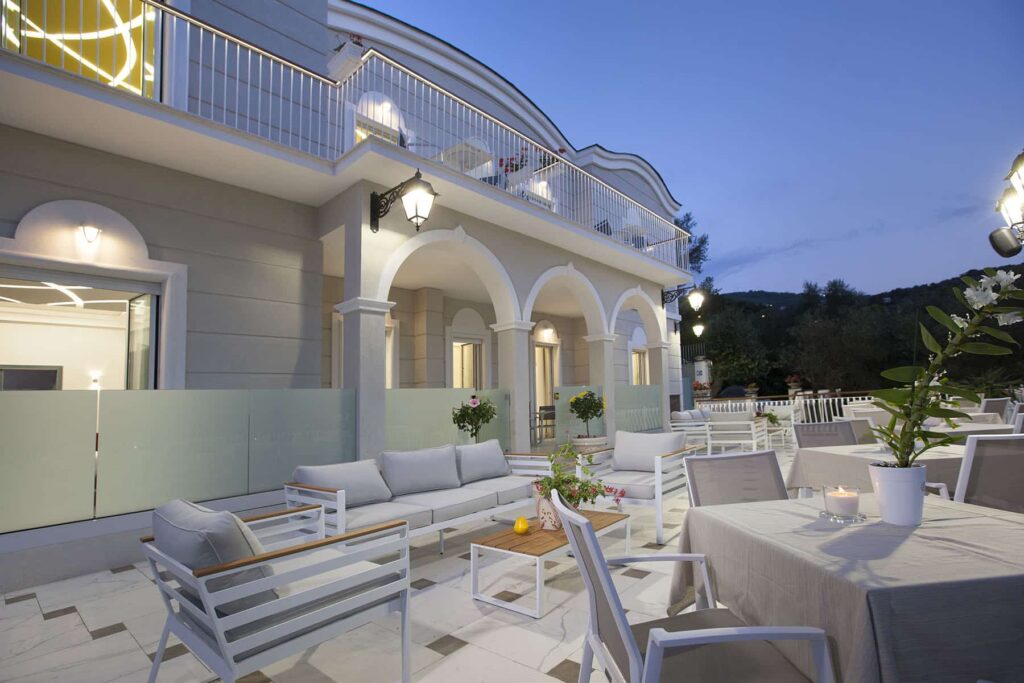 Elegant outdoor seating area of a modern restaurant at dusk, featuring stylish white furniture and soft lighting, surrounded by lush greenery and architectural arches. | Only on SopranoVillas