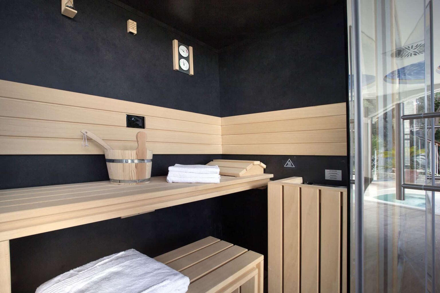 Modern sauna interior featuring wooden benches, a bucket and ladle, neatly folded towels, and a glass door, designed for relaxation and wellness. | Only on SopranoVillas