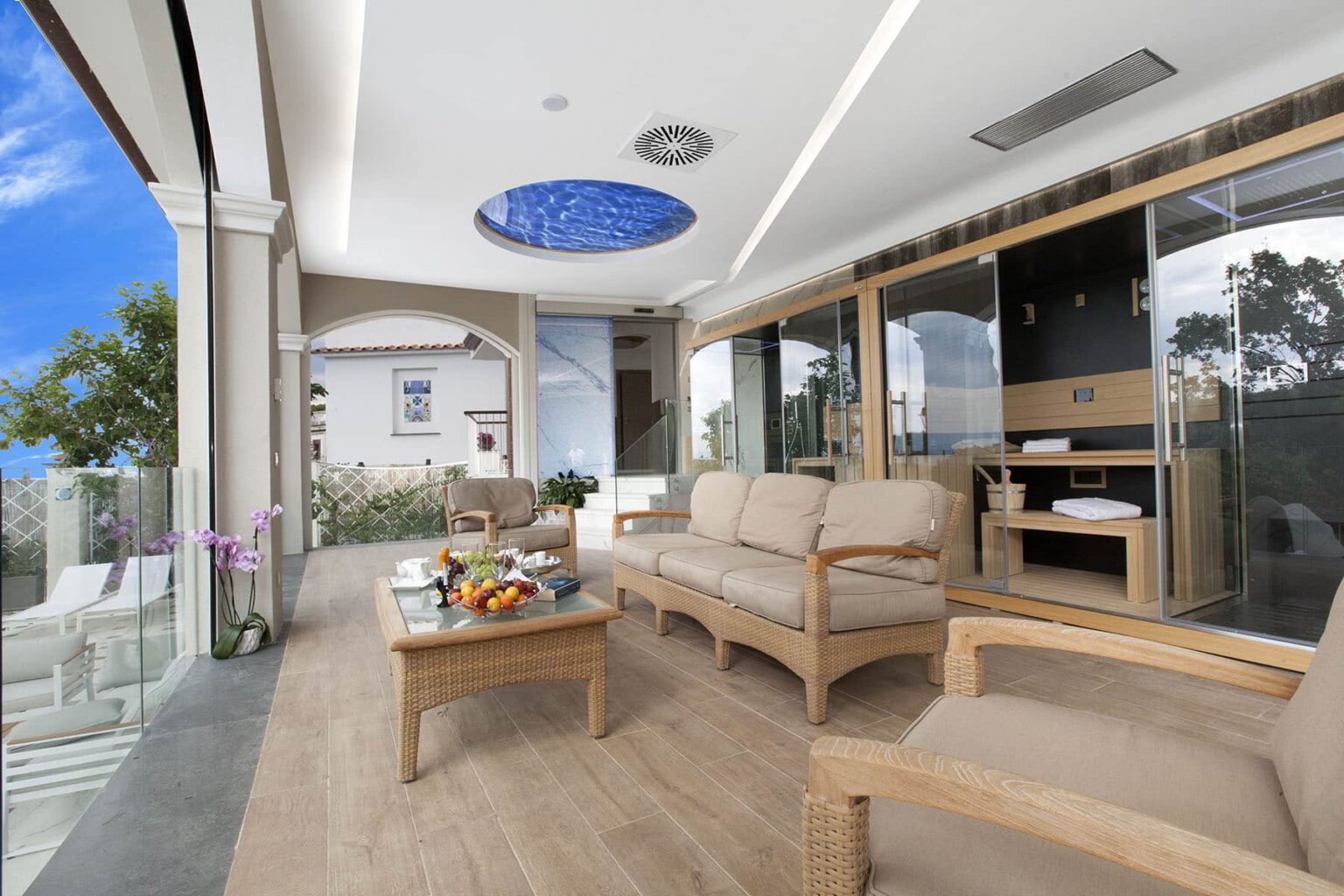 Modern outdoor living space featuring elegant wooden furniture, a decorative ceiling with a blue skylight, and panoramic windows showcasing a scenic view. Ideal for relaxation and entertainment. | Only on SopranoVillas