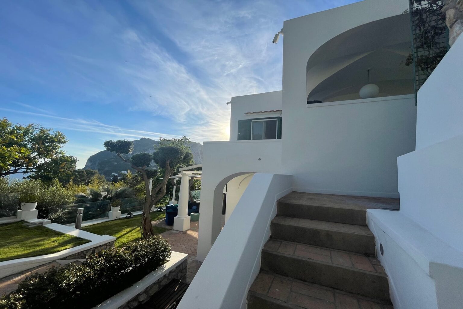 Modern white villa with landscaped garden and stone steps leading to an upper level, set against a clear blue sky. | Only on SopranoVillas