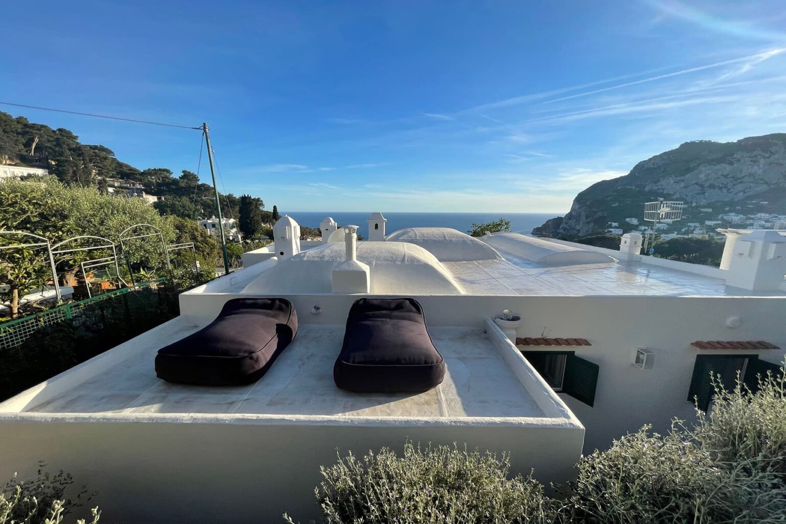 Scenic view from a white rooftop terrace featuring two black lounge chairs, overlooking a coastal landscape with hills and blue sky. | Only on SopranoVillas