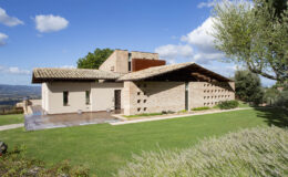 Modern architectural design of a house with a stone facade and a sloped roof, surrounded by green lawn and trees under a clear blue sky.