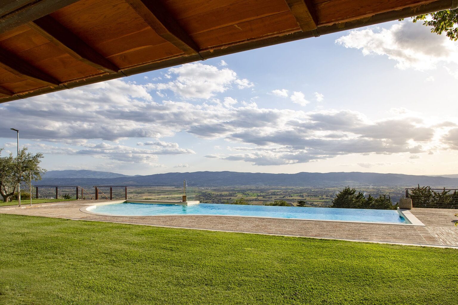 Scenic view of a swimming pool surrounded by lush green grass, with mountains and a cloudy sky in the background, showcasing a tranquil outdoor setting. | Only on SopranoVillas