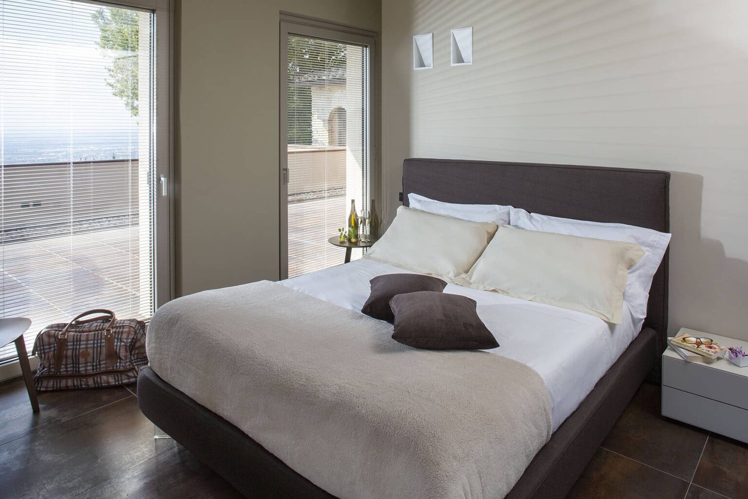 Modern bedroom featuring a comfortable queen-sized bed with neutral bedding, decorative pillows, and a minimalistic design. Natural light streams in through large windows, enhancing the serene atmosphere. The room includes a bedside table and a cozy seating area, ideal for relaxation. | Only on SopranoVillas