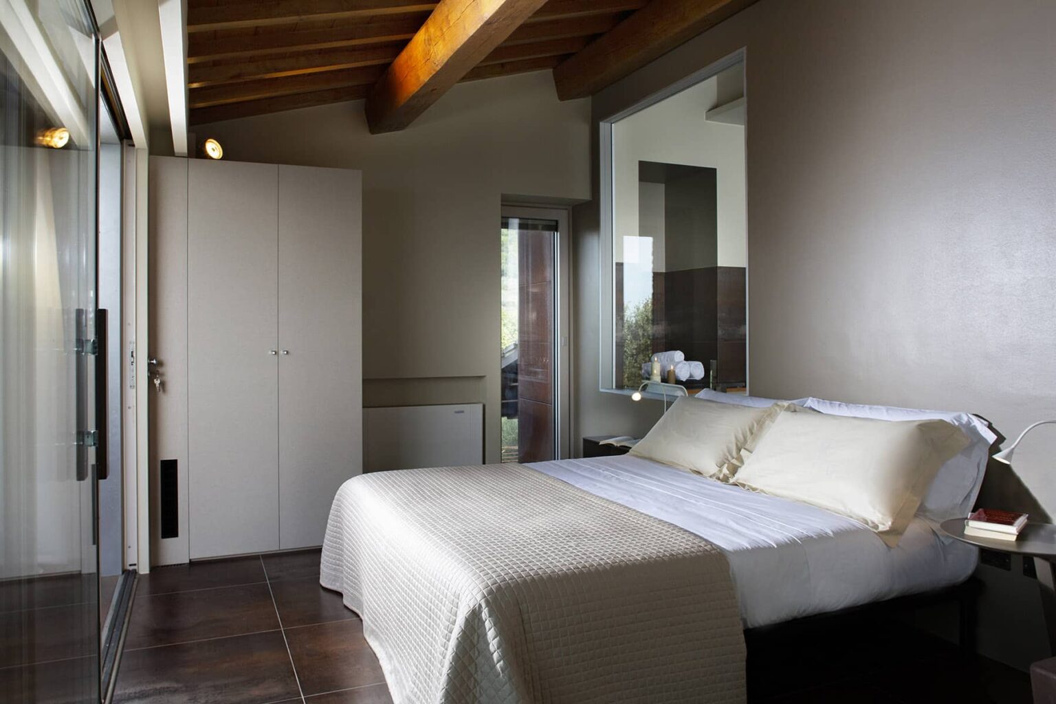 Modern bedroom interior featuring a cozy bed with light-colored bedding, wooden beams, and large windows for natural light. The room includes a sleek wardrobe and minimalist decor, creating a serene atmosphere. | Only on SopranoVillas