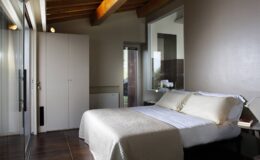 Modern bedroom interior featuring a cozy bed with light-colored bedding, wooden beams, and large windows for natural light. The room includes a sleek wardrobe and minimalist decor, creating a serene atmosphere.