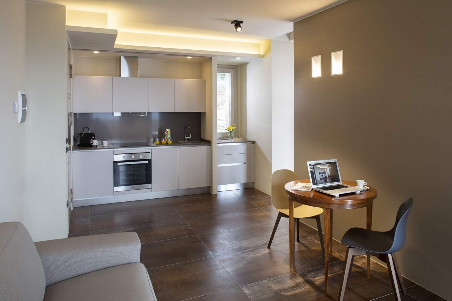 Modern kitchen and dining area featuring a sleek design, equipped with stainless steel appliances, a small dining table with chairs, and natural light from windows, ideal for cozy living spaces. | Only on SopranoVillas