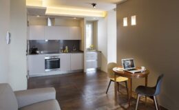 Modern kitchen and dining area featuring a sleek design, equipped with stainless steel appliances, a small dining table with chairs, and natural light from windows, ideal for cozy living spaces.