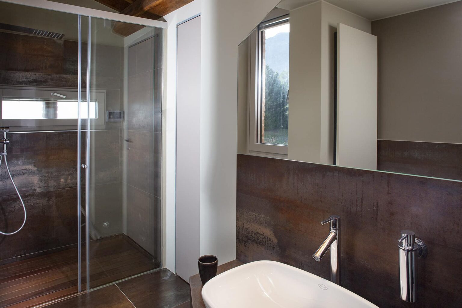 Modern bathroom interior featuring a glass shower enclosure, a freestanding bathtub, and stylish fixtures. The walls are adorned with dark wood accents, and natural light enters through a window, creating a bright and inviting atmosphere. | Only on SopranoVillas
