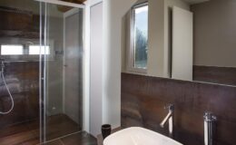 Modern bathroom interior featuring a glass shower enclosure, a freestanding bathtub, and stylish fixtures. The walls are adorned with dark wood accents, and natural light enters through a window, creating a bright and inviting atmosphere.