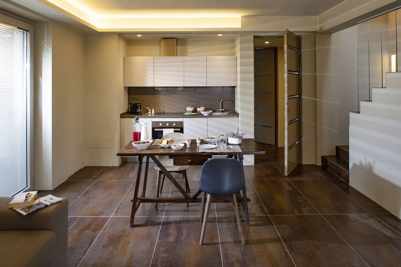 Modern kitchen and dining area featuring a sleek design, wooden flooring, a wooden dining table with a chair, and contemporary cabinetry. Natural light streams through large windows, enhancing the spacious feel. | Only on SopranoVillas