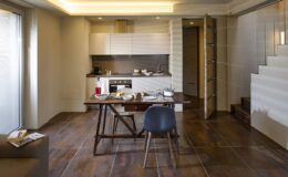 Modern kitchen and dining area featuring a sleek design, wooden flooring, a wooden dining table with a chair, and contemporary cabinetry. Natural light streams through large windows, enhancing the spacious feel.