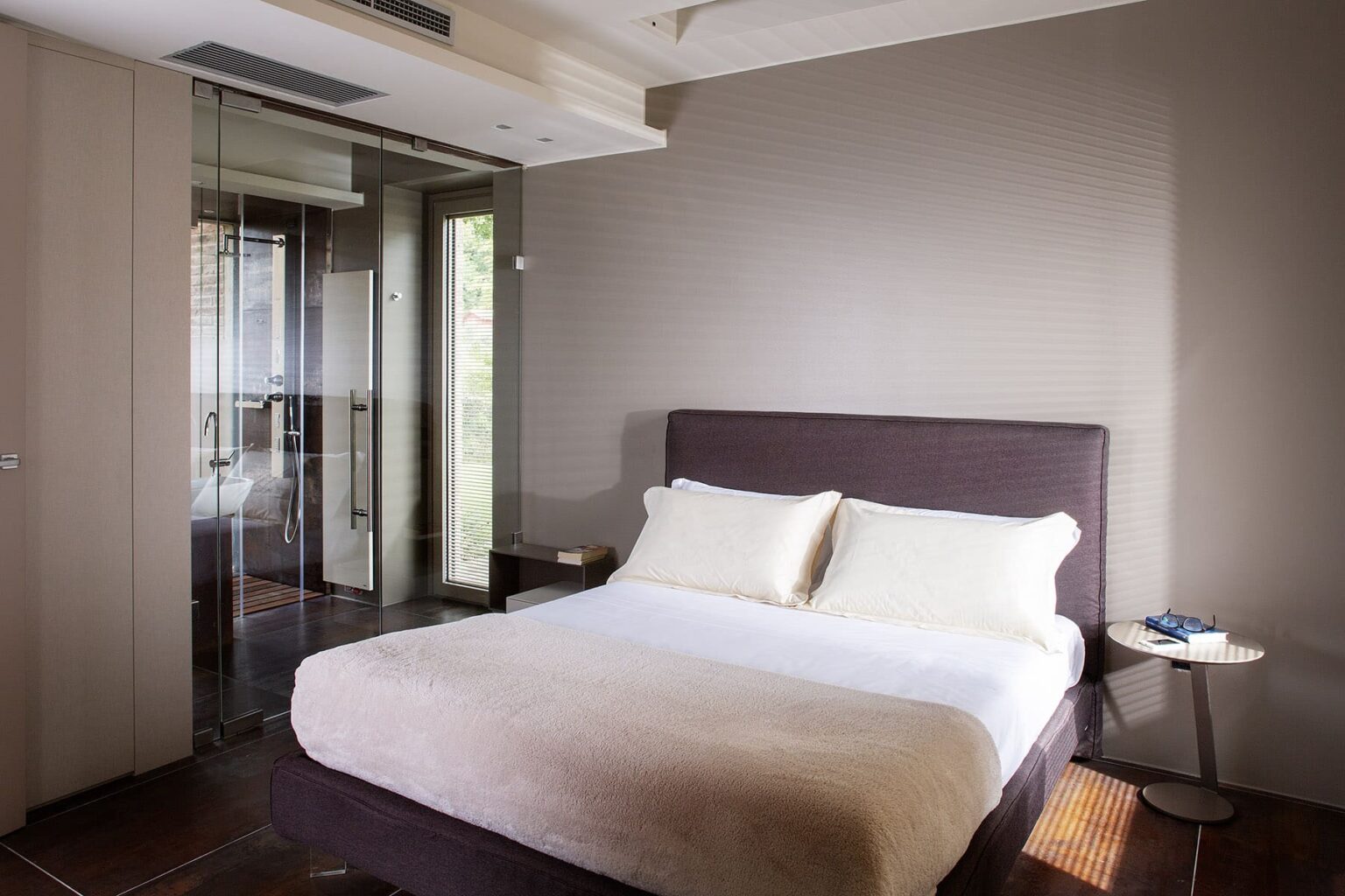Modern bedroom featuring a queen-sized bed with white linens, a bedside table, and large windows providing natural light. The minimalist decor includes soft neutral tones and a sleek glass partition. Ideal for relaxation and comfort. | Only on SopranoVillas