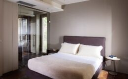 Modern bedroom featuring a queen-sized bed with white linens, a bedside table, and large windows providing natural light. The minimalist decor includes soft neutral tones and a sleek glass partition. Ideal for relaxation and comfort.